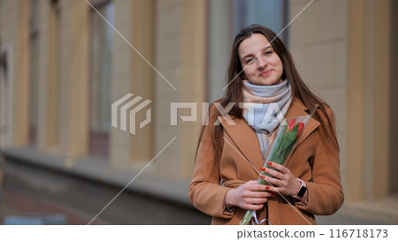 Portrait of a happy brunette girl with a tulip in the background of the city in the cold season. Portrait of a happy brunette girl with a tulip in the background of the city in the cold season. 116718173