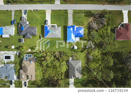 Aerial view of damaged in hurricane Ian house roofs covered with blue protective tarp against rain water leaking until replacement of asphalt shingles Aerial view of damaged in hurricane Ian house roofs covered with blue protective tarp against rain water leaking until replacement of asphalt shingles 116719234