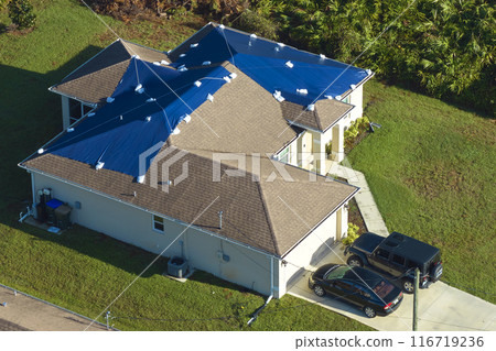 Aerial view of damaged in hurricane Ian house roof covered with blue protective tarp against rain water leaking until replacement of asphalt shingles 116719236