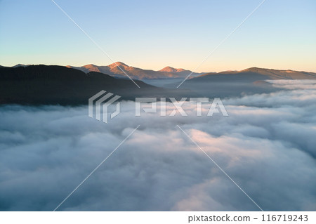 Aerial view of colorful sunrise over white dense fog with distant dark silhouettes of mountain hills on horizon 116719243