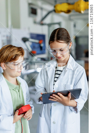 Two classmates standing in robotic laboratory, wearing lab coats and safety eyeglasses. After-school robotics club. Children learning robotics in Elementary school. 116719865