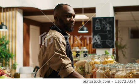 Merchant with apron placing bulk items on shelves at eco store, supporting zero waste concept and sustainable lifestyle. African american vendor advertising homemade pantry supplies. Camera 1. 116720354