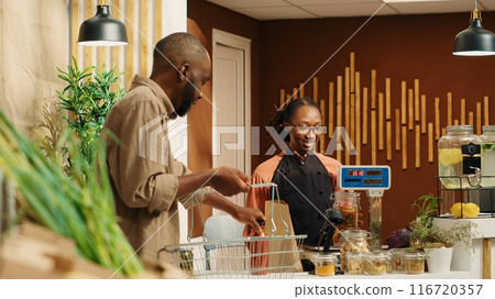 Client giving produce to vendor to weight on local market scale, supporting organic farming and healthy eating. African american merchant scanning goods and bag for buyer at market. Camera 1. 116720357