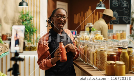 African american vendor promoting homemade sauces and honey, creating online video advertisement to present eco friendly products at zero waste supermarket. Woman recording promotional ad. Camera 1. African american vendor promoting homemade sauces and honey, creating online video advertisement to present eco friendly products at zero waste supermarket. Woman recording promotional ad. Camera 1. 116720358