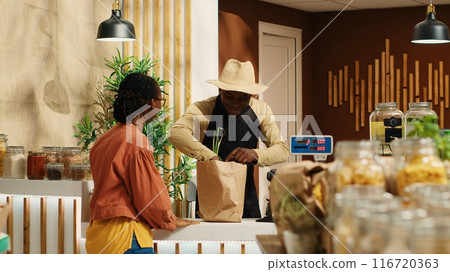 African american vendor weighting produce for regular client at supermarket cash register, using electronic scale to sell locally grown goods. Buyer supporting local farming business. Camera 1. 116720363