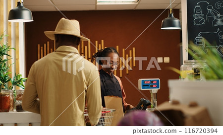 African american seller working at supermarket cash register counter, weighting organic additives free fruits and vegetables. Vendor selling fresh bio produce to male customer at checkout. Camera 1. African american seller working at supermarket cash register counter, weighting organic additives free fruits and vegetables. Vendor selling fresh bio produce to male customer at checkout. Camera 1. 116720394