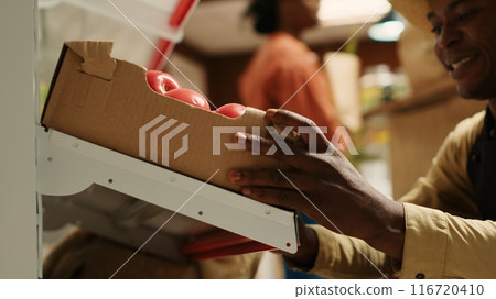 Local farmer placing crates with homegrown produce on shelves, working on fruits and vegetables restock for customers. Merchant carrying boxes of organic freshly harvested goods. Camera 1. 116720410