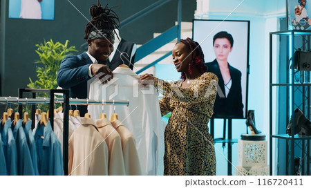 African american worker showing white shirts to pregnant woman, helping her to choose the right size to buy on discount. Store assistant recommending clothes from designer brands. Camera A. African american worker showing white shirts to pregnant woman, helping her to choose the right size to buy on discount. Store assistant recommending clothes from designer brands. Camera A. 116720411