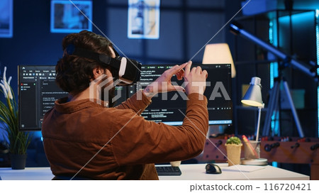 Remote software developer using VR goggles in personal office at home to do javascript programming. IT engineer wearing virtual reality headset in order to visualize lines of code, camera A Remote software developer using VR goggles in personal office at home to do javascript programming. IT engineer wearing virtual reality headset in order to visualize lines of code, camera A 116720421