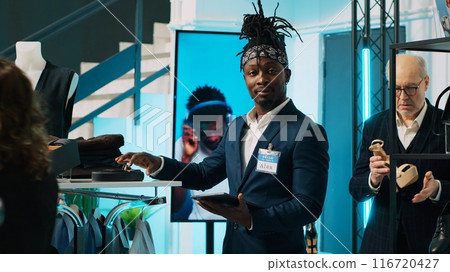 African american worker checking inventory for new clothing line placed on hangers for sale, looking at merchandise on tablet and in store. Retail assistant verifying discount list at mall. Camera A. African american worker checking inventory for new clothing line placed on hangers for sale, looking at merchandise on tablet and in store. Retail assistant verifying discount list at mall. Camera A. 116720427