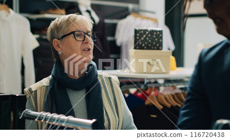 Professional stylist helping senior customer to choose clothes for her new wardrobe, recommending new designer line. Elderly client shopping for formal suit and shirts, retail team. Camera A. Professional stylist helping senior customer to choose clothes for her new wardrobe, recommending new designer line. Elderly client shopping for formal suit and shirts, retail team. Camera A. 116720438