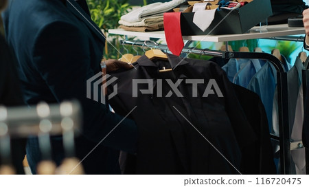 African american worker showing a black shirt and red tie to client, assisting him to buy the perfect outfit for an elegant occasion. Professional stylist gives advice to buyer at store. Camera A. African american worker showing a black shirt and red tie to client, assisting him to buy the perfect outfit for an elegant occasion. Professional stylist gives advice to buyer at store. Camera A. 116720475
