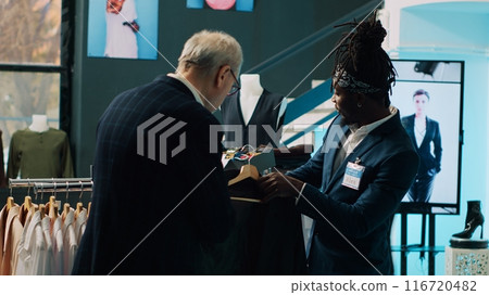 African american store assistant helping regular client with a suit in shopping center, senior customer looking to buy formal clothes. Elderly man shopping for new collection at the mall. Camera A. 116720482