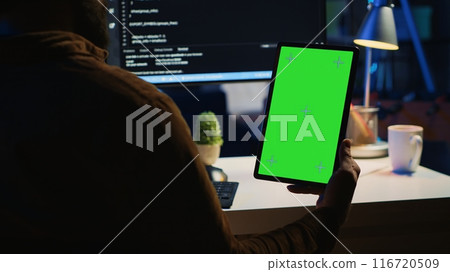Software developer programming in home office, crosschecking lines of code between green screen tablet and PC screen. IT admin holding mockup portable device while coding on computer Software developer programming in home office, crosschecking lines of code between green screen tablet and PC screen. IT admin holding mockup portable device while coding on computer 116720509