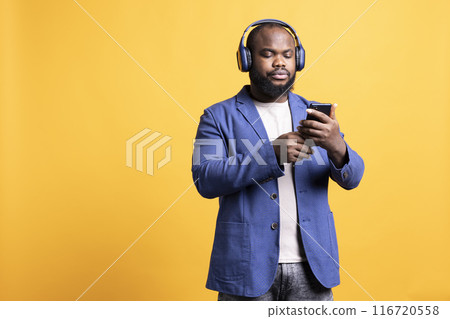 BIPOC man wearing headphones while texting best friend using smartphone. African american person listening music and chatting with mate on mobile phone, isolated over studio background BIPOC man wearing headphones while texting best friend using smartphone. African american person listening music and chatting with mate on mobile phone, isolated over studio background 116720558