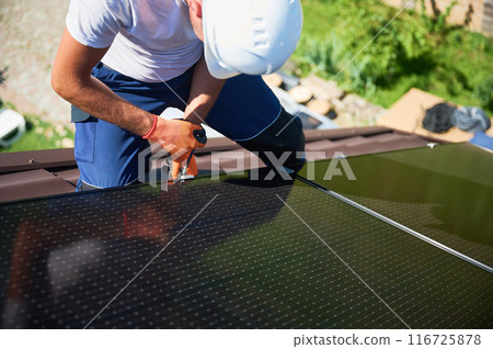 Worker building photovoltaic solar panel system on rooftop of house. Close up of man engineer in helmets and gloves installing solar module with help of hex key outdoors. Alternative, renewable energy 116725878