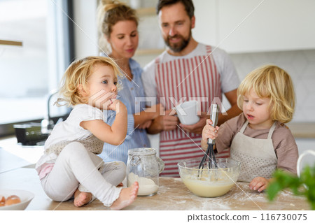 Young nuclear family making pancakes together. Parents and children in kitchen, preparing pancake batter, spending weekend day indoors. Young nuclear family making pancakes together. Parents and children in kitchen, preparing pancake batter, spending weekend day indoors. 116730075