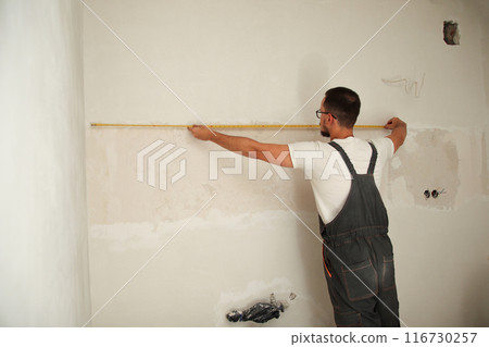 Back view of a construction worker measuring a wall with a tape measure. The skilled builder is using the tape measure on a white wall, highlighting his precision and expertise 116730257