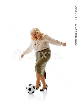 Senior woman, wearing white blouse and green skirt, joyfully kicks soccer ball, looking focused and happy against white studio background. 116735840