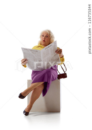 Elderly woman, dressed in yellow blouse and purple skirt, sits on white block, looking surprised while reading newspaper against white studio background. 116735844