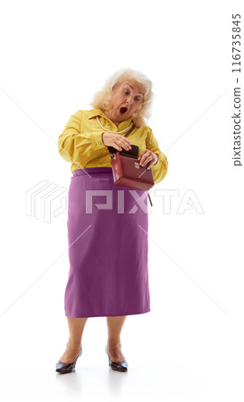 Elderly woman, wearing yellow blouse and purple skirt, looks surprised while peering into her handbag against white studio background. 116735845