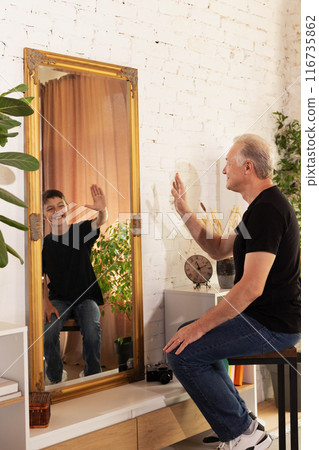 Senior man and his younger self reflected in mirror, both smiling and raising their hands in friendly gesture. Senior man and his younger self reflected in mirror, both smiling and raising their hands in friendly gesture. 116735862