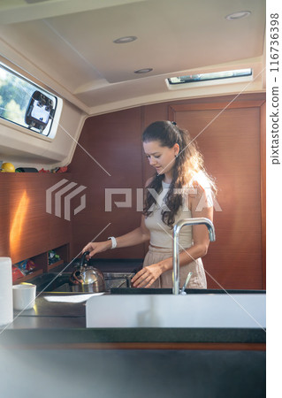Young woman standing and preparing tea in kitchen in cabin of modern yacht during sailing in sea. Yacht life concept 116736398