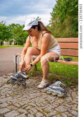 Woman roller skater in the park puts her roller skates on her feet. Outdoor activities. Summer roller skating. Woman roller skater in the park puts her roller skates on her feet. Outdoor activities. Summer roller skating. 116736403