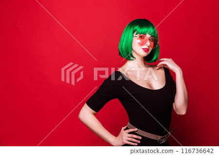 Fashion model posing wearing green wig and sunglasses on red background Fashion model posing wearing green wig and sunglasses on red background 116736642
