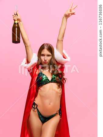 Young woman with slim body, wearing red festive gown and green bikini, standing with champagne bottle and celebrating holiday against pink studio background 116736886
