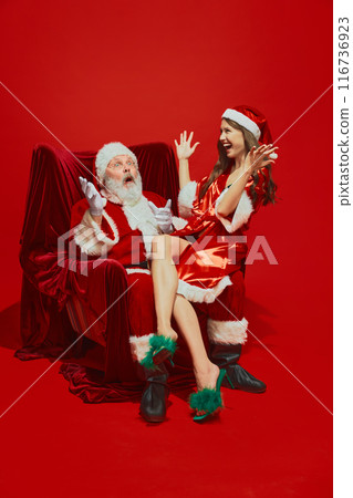 Young woman in red silk gown and heeled shoes sitting on Santa Claus and laughing, telling stories against red studio background 116736923