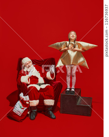 Little girl in star shape costume singing for displeased emotive Santa Claus sitting on chair against red studio background. Performing and expecting gifts Little girl in star shape costume singing for displeased emotive Santa Claus sitting on chair against red studio background. Performing and expecting gifts 116736937