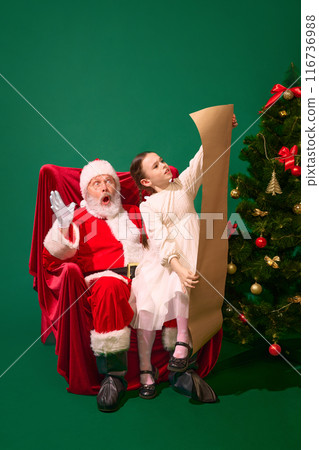Surprised Santa sitting with girl, child reading long wish list beside beautifully decorated Christmas tree, against green studio background Surprised Santa sitting with girl, child reading long wish list beside beautifully decorated Christmas tree, against green studio background 116736988