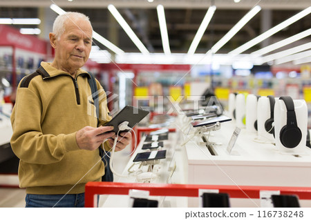 Elderly grayhaired man pensioner examining counter with electronic gadgets and tablets in showroom of digital goods store Elderly grayhaired man pensioner examining counter with electronic gadgets and tablets in showroom of digital goods store 116738248