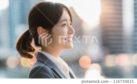A close-up profile of a smiling young woman in a suit with city buildings in the background. 116739815