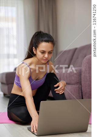 sporty Caucasian woman looking at laptop while sitting on pink fitness mat, ready for online yoga training at home. 116741250