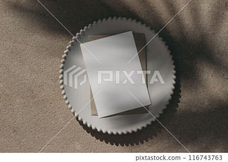 Blank greeting card invitation mockup, envelope on white plate. Beige limestone backgound, palm leaf shadow overlay. Modern template, branding identity. Tropical summer, vacation. Flat lay, top view. 116743763