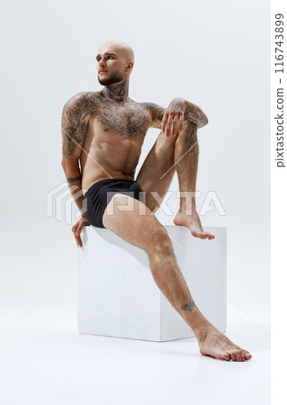Handsome confident young man sitting on cube shirtless, in black underwear, showing strong and athletic body isolated on white studio background 116743899