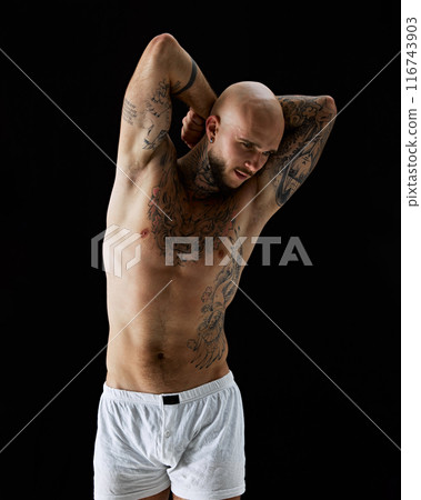 Handsome young guy with fit, relieve, muscular body with tattoos, posing shirtless in white boxers against black studio background Handsome young guy with fit, relieve, muscular body with tattoos, posing shirtless in white boxers against black studio background 116743903