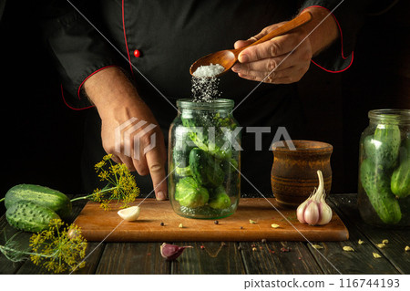 Chef adding salt to a jar of cucumbers before canning. Concept of making delicious pickled cucumbers on the kitchen table by the hands of a cook 116744193