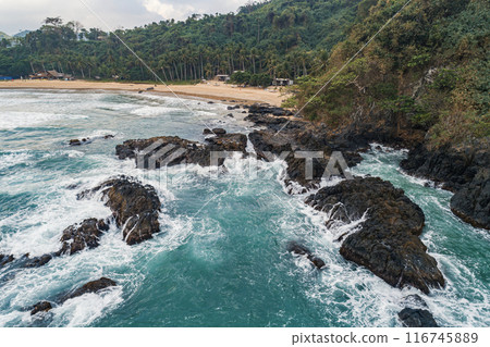 Aerial view of waves crashing on rocky coastline near a tropical beach Aerial view of waves crashing on rocky coastline near a tropical beach 116745889