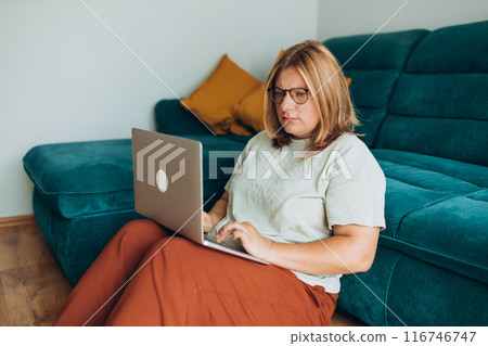 Upset sad, skeptical, serious woman in glasses working at the notebook near the sofa at home. Concepts of education, work and comfort. Negative people emotion, pensive girl sitting near sofa 116746747