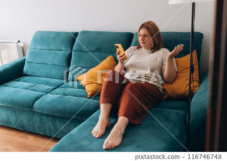 Upset sad, skeptical, serious woman holding smartphone gadget for communicate indoors. Negative people emotion, pensive girl sitting at home. Redhead woman holding smartphone. Screaming, hate, rage 116746748