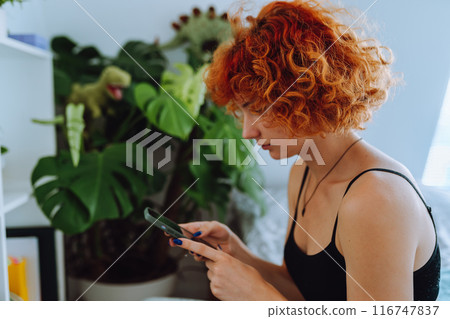 portrait of young woman with curly red hair sitting on bed in bedroom using phone 116747837