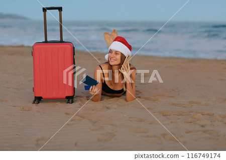 Happy young woman in Christmas Santa Claus hat lying on sand seashore with red suitcase holds smartphone, credit card and passport looks at camera. New Year winter holiday vacation at tropical resort Happy young woman in Christmas Santa Claus hat lying on sand seashore with red suitcase holds smartphone, credit card and passport looks at camera. New Year winter holiday vacation at tropical resort 116749174