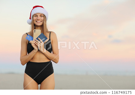 Happy slim young woman in Santa Claus hat, bikini outdoors shows smartphone, credit card, passport for book online ticket to travel on Christmas and New Year 2024 holiday vacation at tropical resort Happy slim young woman in Santa Claus hat, bikini outdoors shows smartphone, credit card, passport for book online ticket to travel on Christmas and New Year 2024 holiday vacation at tropical resort 116749175