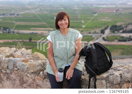 Active middle aged woman with backpack relaxes sits with water bottle on hill, looks at beautiful view after hiking up. Healthy lifestyle in retirement. Adventure seeks woman treks enjoys freedom 116749185