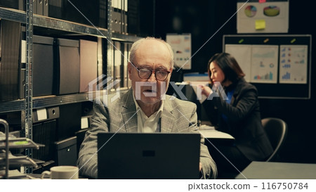 Senior law agent examining financial records to find account information and find criminal, working with forensic expert in incident room. Police department investigation. Camera B. 116750784