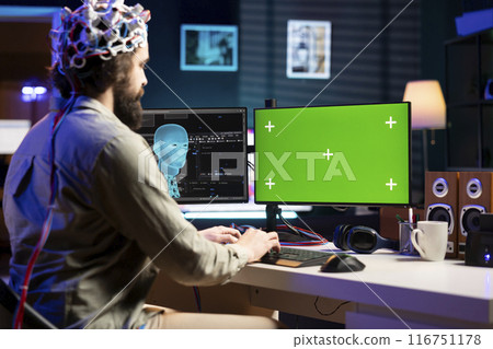 IT technician using EEG headset device translating thoughts into commands on green screen PC display on desk. System administrator controlling chroma key computer functions using mind 116751178