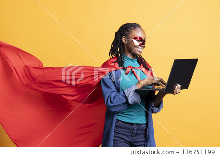 Happy girl portraying superhero with cape hiding identity, working on laptop, studio background. Woman wearing cloak posing as hero in costume, typing on notebook keyboard, covering face with mask Happy girl portraying superhero with cape hiding identity, working on laptop, studio background. Woman wearing cloak posing as hero in costume, typing on notebook keyboard, covering face with mask 116751199